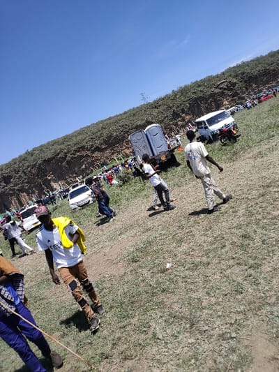 The image displays a large outdoor gathering under a clear blue sky in Olkaria ward, Kenya. The scene is set on an expansive, uneven field consisting of dry, light brown earth with sparse patches of green grass. In the background, a prominent, vegetated rocky escarpment dominates the horizon, with a power transmission line and pylon visible near its crest.
Numerous vehicles, including several white sedans, a white van, and other cars of various colors, are parked or positioned further back on the field, near the base of the escarpment. A significant crowd of people is congregated in this mid-ground area. Centrally, two grey portable toilets are secured on the bed of a dark flatbed truck or trailer. Adjacent to this setup, a white van and a red motorcycle are parked.
In the immediate foreground, several individuals are walking across the field. One male subject, positioned toward the left, is wearing a white t-shirt with a dark logo, brown ripped jeans with black knee patches, a maroon baseball cap, and a yellow jacket draped over his left shoulder. He is walking diagonally towards the lower left of the frame. Other individuals are observed walking towards the right, dressed in casual attire such as t-shirts and pants. No direct interactions between individuals in the foreground are clearly discernible. The overall environment suggests a sunny day at a public event or gathering.