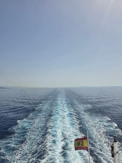 The image depicts a wide expanse of the Mediterranean Sea, viewed from the stern of a vessel. The water is a deep blue, disturbed by the white, frothy wake left by the ship. In the foreground, a Spanish flag, red and yellow with the letters "M9" in black, flies from a white flagpole. To the right, a white pole with some nautical equipment is visible. The horizon is a faint line where the sea meets the sky, with distant landmasses or islands visible on the left and in the far distance. The sky is a pale, clear blue with a subtle haze near the horizon and a bright sun flare in the upper right corner. The scene is tranquil and suggests travel or passage through open waters. The location is identified as Santa Ponça, Spain, which is a coastal town on the island of Mallorca, known for its bay and marina.
