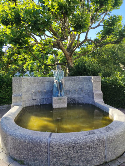 A statue of a seated figure holding a spear is positioned behind a fountain in the city of Achern, Germany. The fountain has a curved basin made of speckled gray stone, filled with yellowish water. Behind the basin, a stone wall with three spouts is visible, from which water flows into the basin. The statue, cast in a greenish-blue metal, sits on a stone pedestal in front of the wall. Above the fountain, a large tree with lush green leaves dominates the upper portion of the image, with sunlight filtering through its branches. Dense green foliage forms a backdrop on the right side of the scene. The ground around the fountain is paved with light-colored bricks.