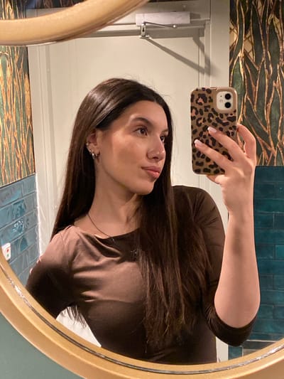 A young woman takes a selfie in a bathroom mirror. The bathroom has a teal tile backsplash, patterned wallpaper, and white molding. The woman is wearing a brown long-sleeved shirt and has dark hair. She is holding a phone with a leopard print case. The location is likely in Uppsala, Sweden, as the prompt specifies.