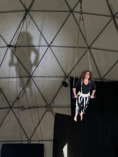 A medium shot shows a female aerialist performing in what appears to be a circus tent. The aerialist has curly brown hair, is wearing a black, long-sleeved top, and black-and-white striped pants. She is hanging from a trapeze swing constructed of rope and what appears to be a blue padded seat. She is barefoot. The tent's structure is visible behind her, a geodesic dome of white panels sectioned by metal struts. Electrical cords and equipment are visible attached to the structure. A black curtain is partially visible in the background to the right. The location is Menashe Regional Council, Israel.