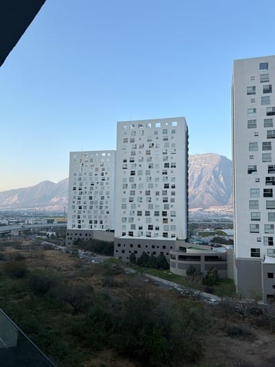 Three white, high-rise buildings stand vertically against a blue sky. Each building has numerous square windows. In the background is a large, mountainous range. Dense vegetation is visible on the right side of the image, suggesting a hillside or park area. An urban landscape stretches between the buildings and the mountain.