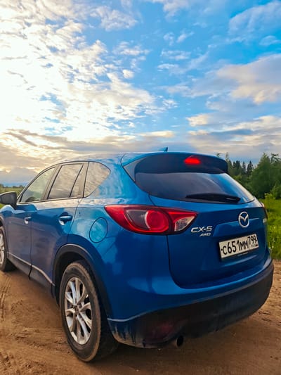 A blue Mazda CX-5 AWD is parked on a dirt road in what appears to be a rural or semi-rural area. The vehicle is facing away from the viewer and the rear passenger side is visible. The license plate reads "C651MM799 RUS". The background shows a field and trees, with a partly cloudy sky above. The scene is located in Курово, Russia.