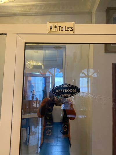 The image shows a glass door with a sign that reads "RESTROOM" in black lettering on a gold background. The sign is attached to the top of the door with a white frame around it. Above the door, there is another sign that reads "Toilets" in white lettering on a black background. The sign depicts a stick figure of a man and a woman. The door is partially open and the interior is visible. Inside the door, a woman is reflected in the glass. The woman is wearing an orange jacket and a black sash with gold lettering that says "LIFE FOR 2121". The woman is looking towards the camera. The environment is a modern interior. The image is taken from the outside looking in at the door. The location of the image is Buea, Cameroon at latitude: 4.145, longitude: 9.23.