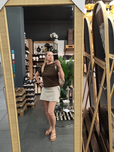 A woman is captured taking a mirror selfie in what appears to be an indoor retail space, likely a home decor or gift shop. She stands with one hand on her hip, holding a smartphone in her other hand to capture her reflection in a large, rectangular mirror framed with a light, woven bamboo-like material.
The woman has light-colored hair tied back in a ponytail and is wearing a dark brown, ribbed short-sleeve top with a subtle animal print pattern, a light-colored, possibly cream or beige, mini skirt, and tan flat sandals with straps. A light-colored, textured handbag is slung over her shoulder, and she wears a smartwatch on her left wrist. Her toenails are painted a dark color.
The background, visible through the mirror, reveals a commercial setting. To her left, against a dark gray wall, are wooden shelving units displaying various decorative items including white ceramic busts or heads, black and gold ornate wall plates, candles, and other packaged goods in cardboard boxes or crates on the floor. An artificial orchid plant and a green leafy plant add to the decor. To her right, a glass coffee table with a shaker bottle and a black and white striped rug underneath is visible. Further right, multiple standalone mirrors with arched tops and wooden frames are propped up. The floor is covered in large, dark gray tiles. The overall ambiance suggests a modern and stylish retail environment.
The lighting indicates daytime. No legible text is visible, although small colored rectangles and faint labels can be seen on shelves and the wall. The woman's expression is neutral and focused on her phone.