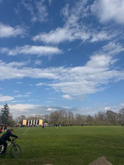 The image depicts an outdoor scene likely in or near a park or recreation field. The dominant feature on the left side is a wide expanse of blue sky dotted with elongated, streaky white clouds. The right side of the image primarily features a green, grassy field, with a treeline visible in the distance. A building structure is visible in the midground, appearing to be made of light-colored material. A number of individuals are visible along the periphery of the field, suggesting an event or gathering is taking place. In the foreground, a person is holding a bicycle. The location in Gromshin, Bulgaria is not evident from the image.