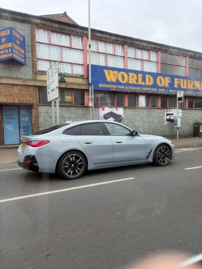 The image presents an overhead street view capturing a light grey modern sedan driving on an asphalt road next to a commercial building in Leicester, United Kingdom, under an overcast sky. The car, featuring multi-spoke wheels with black and silver accents and visible red brake calipers on the front, is moving from right to left in the frame.
The multi-story building alongside the road appears to be a furniture store. A large blue sign prominently displays "WORLD OF FURNITURE" and lists offerings such as "REPRODUCTION & ITALIAN FURNITURE, BEDS, 3 PIECE SUITES, CARPETS, RECLINERS." A smaller blue sign further down advertises "WORLD OF FURNITURE" with a "20,000 Sq ft SHOWROOM." Upper windows are numerous, some covered by white curtains, and a "SALE 50% OFF" sign is visible in one. The lower part of the building has brick and stone cladding, with two blue doors covered in graffiti.
On the pavement between the road and the building, a white street sign with directional arrows points towards locations like "Ring Road", "North", "South", "Park", and "Shopping." A bus stop sign and a waste bin bearing the "Virgin" logo are also present. The road features clear white lane markings. The car's rear license plate is partially visible, beginning with "Y7". The scene portrays a daytime urban setting.