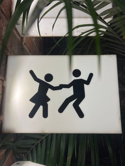 The image features a brightly illuminated rectangular sign displaying two stylized human figures in black silhouette. The upper figure, resembling a man, is inverted, balancing on one hand with his other arm outstretched and legs dynamically posed. The lower figure, resembling a woman in a skirt, is upright, reaching up to grasp the man's hand. Their combined pose suggests a playful, acrobatic dance, or a dynamic interaction.

The sign glows with a soft white light, standing out against a dark, shadowy background. Lush, green, palm-like foliage partially frames the sign, with leaves in the foreground slightly blurring the view. On the far left, another similar, partially visible white rectangular object also emits light. A reddish-brown brick surface is visible at the bottom of the frame, serving as a base or ground.

The strong illumination of the sign against the darkness indicates it is nighttime or located in a dimly lit indoor space. The tropical vegetation and brickwork, combined with the location context of Mérida, Mexico, suggest an outdoor or semi-outdoor setting such as a garden, courtyard, or patio of a restaurant, bar, or other establishment. The whimsical nature of the figures implies a fun or unique atmosphere, possibly indicating a restroom or a themed area, without relying on traditional text.