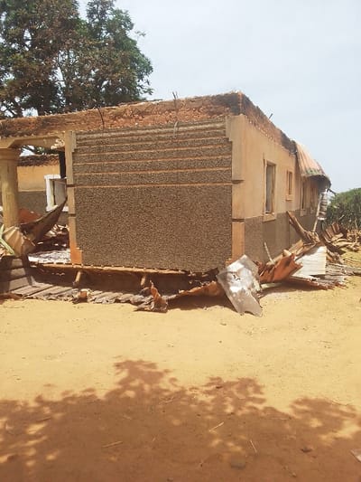 The image shows the remains of a building in Kwal, Nigeria, at the coordinates 9.804° N, 8.647° E. The building is a one-story structure with a concrete foundation and a corrugated metal roof. The roof has been partially destroyed, and some of the walls have collapsed. The building appears to have been damaged by fire. There is debris scattered around the building, including pieces of metal and wood. The ground in front of the building is dirt, and there is a tree in the background. There are no people visible in the image.