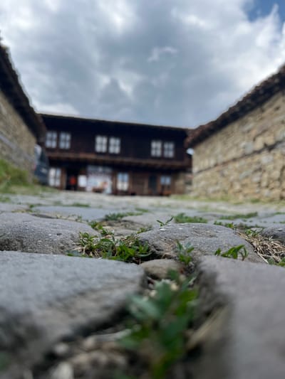 The image is a low-angle, close-up shot of cobblestones with small green plants growing between them, set against a blurred background of traditional Bulgarian architecture in Zheravna, Bulgaria. The foreground is dominated by large, gray cobblestones, some of which are slightly damp. Small sprigs of green grass and weeds emerge from the cracks and crevices of the stones.
In the mid-ground, a building constructed with dark wood and stone is visible. It features multiple windows with white panes, arranged in symmetrical patterns on two levels. The roof has dark tiles. To the left and right of this central building, other stone structures with similar tiled roofs frame the scene, creating a sense of a courtyard or village street. The sky above is overcast with heavy, gray clouds, suggesting a subdued or impending weather change. There are no discernible people or animals in the frame. The overall impression is of a rustic, historic setting captured from a ground-level perspective.