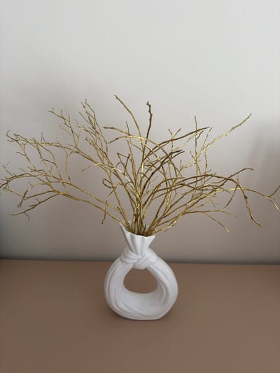 The image shows a white, twisted vase with gold branches inside, placed on a brown surface in front of a beige wall. The vase has a unique, knot-like design at its opening, where the gold branches emerge. The branches are thin and spread out, some reaching upwards and outwards. The background is a plain, textured beige wall, and the surface below is a smooth, matte brown. The lighting is soft, casting subtle shadows. The scene is static and appears to be an indoor setting, possibly a home or studio. There are no people or animals present.