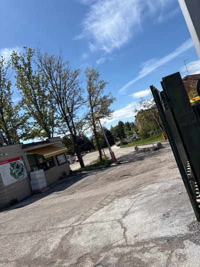 The image captures an outdoor scene under a clear, bright blue sky dotted with wispy white clouds, suggesting a sunny day. The perspective appears to be slightly upward, looking through an opening.

In the foreground, a dark green or black heavy-duty overhead structure or gate stretches across the top of the frame. Below this, a paved road or driveway, marked with numerous cracks and patches, extends into the middle distance on the right.

To the left, a light-colored, possibly concrete or stone building with a yellow awning is visible. Attached to the building is a large informational sign featuring a map of a complex. Visible text on this sign includes "COMPLESSO DI COLLE PARADISO" (Complex of Colle Paradiso) and "ADIS MARCHE," along with labels for "Campus 1," "Campus 2," "Campus 3," etc., outlining several buildings. Near the building, there are some potted plants and what appear to be stacks of white, shrink-wrapped boxes.

A long, white and orange barrier arm, typically used for controlling vehicle access, is extended across the paved area in the middle ground. Further down the road, another lamppost and more trees line the path. Numerous trees with fresh green leaves, indicative of spring or summer, are scattered throughout the scene, particularly on the left and in the background, contrasting with the bare branches higher up.

The scene depicts a static outdoor environment, possibly an entrance to a university campus or institutional complex, given the "Colle Paradiso" and "Campus" references, consistent with the Camerino, Italy location context. No people are visible, and there is no specific activity taking place. The time of day appears to be daylight, likely morning or afternoon, due to the bright sun and clear sky.
