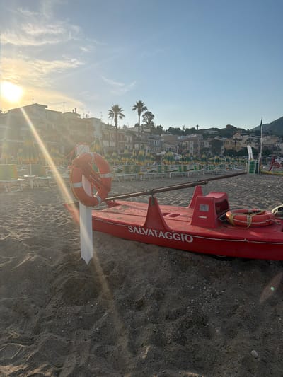 The scene is a beach in Chianchitta, Italy. A red lifeguard boat, labeled "SALVATAGGIO," rests on the sand, with a life preserver affixed to it. Beach umbrellas and chairs are visible in the background, near buildings and palm trees. The sky is blue with some clouds, and the sun is low, creating lens flare. The sand is dark and textured.