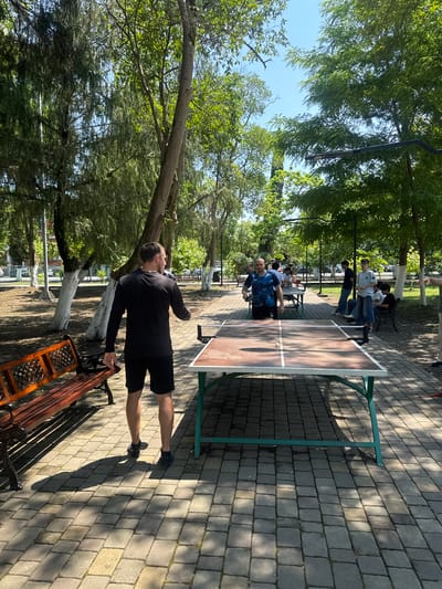 A man in a black long-sleeved shirt and black shorts stands with his back to the camera, facing a ping pong table. He is in the foreground on the left side of the image, with his right arm extended as if to hit a ball. A ping pong ball is visible on the table. In the background, several other people are gathered around other ping pong tables, some playing and some spectating. The scene takes place outdoors on a paved area with trees and benches. This is likely in Poti, Georgia, where outdoor recreational facilities are common.
