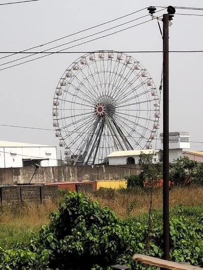 The image displays a large, stationary Ferris wheel in Magado, Nigeria, under an overcast, light grey sky. The Ferris wheel dominates the upper-middle frame, featuring a silver-colored structural framework with numerous spokes radiating from a central hub adorned with a red and white starburst design. Multiple light-colored gondolas with red trim are evenly spaced around its circumference.
In the foreground, dense green foliage occupies the lower left, transitioning to tall, dry, yellowish-brown grass and weeds across the lower midground. A dark brown utility pole with three horizontal power lines extends vertically along the right side of the frame. A horizontal wooden beam is partially visible at the bottom right.
Behind the foreground vegetation, a concrete wall runs horizontally. Structures visible beyond this wall include a large, white industrial-style building with a flat roof on the left, and a smaller white building with a light-colored, sloped roof on the right. Directly behind the concrete wall, an orange-red shipping container and a yellow shipping container are positioned. No individuals are present in the scene.