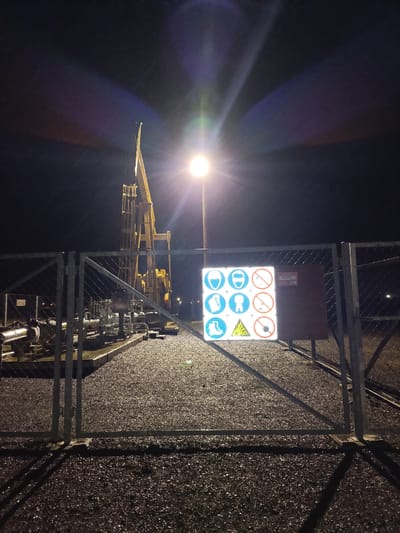 The image depicts an outdoor industrial scene at night, illuminated by a bright overhead light that creates lens flare. In the background, a yellow oil pumpjack stands against a dark sky. In the foreground, a chain-link fence with a gate separates the viewer from the industrial equipment. Attached to the fence is a square sign with multiple circular safety symbols, including representations of a hard hat, safety glasses, hearing protection, gloves, protective clothing, and safety boots. There are also prohibition symbols indicating no smoking, no open flames, and no bare hands. To the right of the safety sign is a reddish-brown metal box with some writing on it. The ground is covered with dark gravel. There are no people visible in the image. The location is identified as being in the city of Unknown, Kazakhstan.