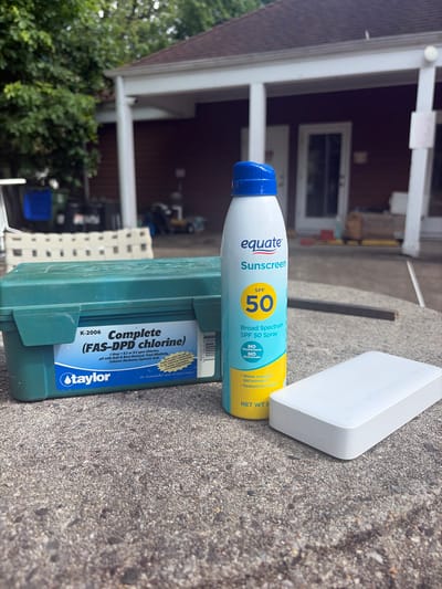A slightly high-angle, close-up outdoor shot shows an assortment of objects resting on a gray concrete surface. These include a green Taylor K-2006 Complete (FAS-DPD chlorine) water testing kit, a yellow and light blue Equate SPF 50 sunscreen spray bottle, and a white, rectangular electronic device, possibly a portable charger, situated behind the sunscreen bottle. The objects appear to be haphazardly placed, suggesting a temporary arrangement.
In the blurred background, a portion of a two-story building with white support pillars and a dark-colored exterior is visible. Some greenery, possibly trees or shrubs, flanks the left side of the building, suggesting a residential or suburban setting. Other details, such as a ladder and a blue container, are discernible in the background.
While the image offers clues about the setting and objects, a precise location within Edison, United States of America, cannot be definitively determined without additional information or context.