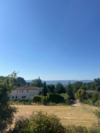 The image depicts a rural landscape under a clear, bright blue sky in Puyloubier, France. In the foreground, a field of dry, light-brown grass occupies the lower portion, with green shrubs and bushes partially obscuring the bottom edge. In the midground, towards the left, a multi-story, light-colored house with a red-tiled roof is visible, featuring multiple windows, some with dark shutters. The house is partially framed by dense green foliage. A variety of trees, including tall, slender conifers and broadleaf trees, populate the area around the house and extend across the midground. A narrow, unpaved path winds through the vegetation on the right side of the midground. The background features distant, hazy blue-grey mountains or hills stretching across the horizon. No individuals, specific objects beyond natural elements and the single structure, or discernible actions are present within the scene. The lighting suggests daylight conditions.