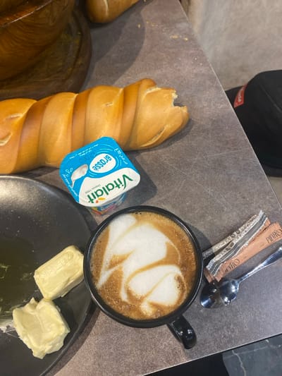 An overhead close-up view presents a breakfast or snack arrangement on a dark grey, textured table, located in the city of Monastir, Tunisia. The primary subjects include a golden-brown, braided loaf of bread, partially consumed with a visible bite mark on one end. Adjacent to the bread is a small, light blue and white plastic cup of Vitalait Brasse yogurt. A black mug containing a cappuccino or latte, featuring intricate latte art in the foam, is positioned to the right. On a dark grey plate to the left, two distinct rectangular pats of butter are visible. A silver spoon and two sugar packets, one brown and one patterned, both branded "PIEVRO," are placed next to the coffee mug. The background shows out-of-focus elements, including a section of a wooden bowl, another bread item, and a dark object with a red tag. No individuals are depicted.