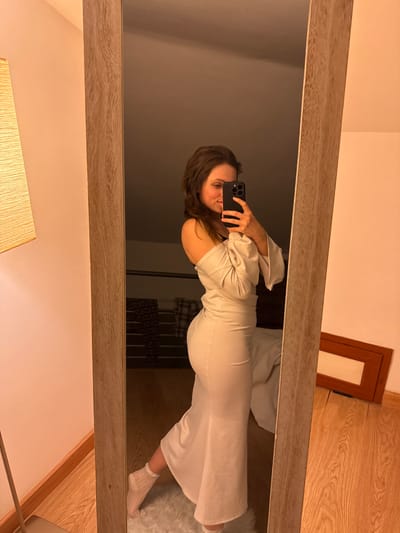A female individual is captured in a mirror selfie within an indoor setting in Cava de' Tirreni, Italy. She wears a light-colored, off-the-shoulder dress with long sleeves, form-fitting around the torso and hips, flaring near the calves, and white frilly socks. A black bra strap is visible on her left shoulder. The subject holds a dark smartphone with both hands, aiming it towards the mirror, with a slight smile directed at the phone's screen. The mirror features a light wooden frame. Reflected within the mirror, behind the individual, is a warm-toned lamp with a rectangular fabric shade, dark patterned clothing folded on a surface, and a light-colored bedspread. A white shaggy rug is underfoot, with a wooden floor visible beyond. A brown rectangular object, likely a frame, lies on the wooden floor to the right of the reflected scene. The room's ambient lighting is warm, and the wall opposite the mirror is a light pinkish-beige with a wooden baseboard.