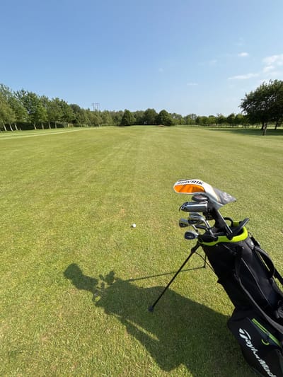 The image shows a golf course in Pocklington, UK. It features a golf bag with clubs standing on a grassy fairway, with a single golf ball lying on the green grass. The bag is black with a green and yellow logo that reads "TaylorMade." The clubs have black shafts with a white driver head featuring the logo "MAVRIK." The background is a bright blue sky with a few white clouds, and the image shows lush green grass covering the ground. The fairway has a gentle curve, with a small copse of trees on the far side of the fairway. The golf ball lies a few feet in front of the bag. There are no people present in the image. The image is taken from a low angle, likely from the perspective of a golfer. The image shows a typical golf course scene, with an emphasis on the golfer's equipment and their position on the fairway.