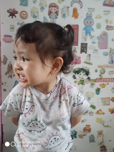 A young child with dark hair pulled back in a small ponytail looks to the side with a slight smile, revealing their teeth. The child is wearing a white t-shirt patterned with dinosaurs. The background is a wall covered in colorful cartoon stickers of various characters and objects, adding a playful and whimsical element to the scene. The lower left corner has a watermark indicating the photo was taken on a Redmi 7 AI Dual Camera.