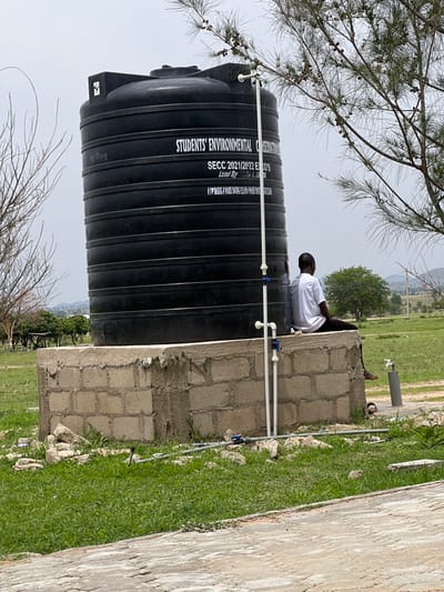 The image, taken in Yali, Nigeria, features a large black water tank prominently in the foreground. The tank has text printed on it, including "STUDENTS' ENVIRONMENTAL O," "SECC 2021/2022 ECO'S," and "Lead By A. BABA." A person in a white shirt is sitting on the edge of a concrete structure next to the water tank, facing away from the camera. Behind them, a grassy area stretches out with some trees and hills visible in the distance, suggesting a rural or semi-rural setting. The weather appears to be overcast or hazy. The image is oriented vertically, with the water tank taking up a significant portion of the left side. The scene depicts a moment of rest or observation by the individual next to the essential water storage infrastructure.