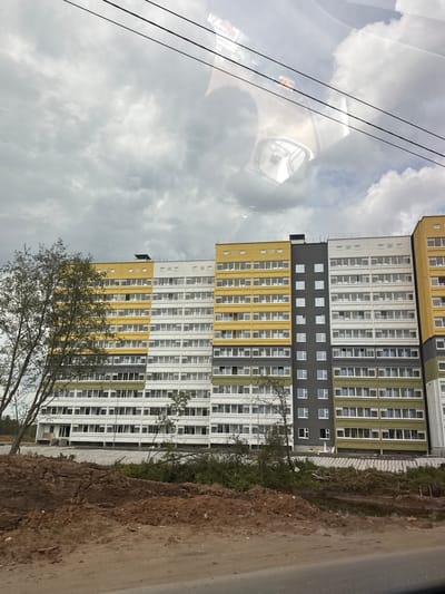 The image captures a large, multi-story residential building, likely under construction or recently completed, viewed from inside a vehicle. The perspective is through a window, as evidenced by prominent reflections on the left side.

The building is imposing, composed of several contiguous blocks with varying facade colors including yellow, white, dark grey, and olive green, suggesting a modern architectural style. It features numerous rectangular windows uniformly arranged across its many floors. While largely complete, some sections or surrounding areas appear to be still under development.

In the foreground, a tree with sparse foliage partially obscures the building on the left. Along the base of the building on the right, a large mound of disturbed earth or construction debris is visible, next to what appears to be a dirt road or pathway. The sky above is heavily overcast, filled with white and grey clouds, indicating a cloudy daytime setting.

The reflections on the car window on the left side of the frame include distorted views of the cloudy sky, thin power lines stretching horizontally, and elements of the car's interior, notably a side mirror and parts of the dashboard or console. No people or visible text are present. The scene depicts an urban or suburban development in Kabanikha, Russia, under an overcast sky.