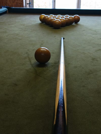 A low-angle, eye-level perspective captures a billiard game setup in an indoor setting located in Терновский округ, Ukraine. A billiard table, covered in a worn olive-green felt, dominates the frame. In the foreground, slightly left of center, a single solid brown ball rests on the felt. Extending from the bottom center of the frame and angled slightly towards the left is a wooden billiard cue stick, featuring a light wood shaft and a darker, stained grip section. Its tip is positioned to strike the brown ball. In the mid-ground, at the far end of the table and positioned directly in line with the cue stick, a complete rack of fifteen identical solid brown billiard balls is tightly arranged in a triangular formation. The background features large, out-of-focus windows, through which bright daylight illuminates the scene, casting subtle reflections on the balls and cue. A dark green or black table rail is visible along the top edge of the table. No individuals are present, but the setup suggests an imminent shot in a game of billiards.