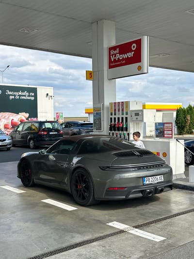 A grey Porsche 911 Targa 4 GTS with Bulgarian license plates PB 2206 XE is parked at a Shell V-Power gas station in Drazhevo, Bulgaria. A young boy in a white t-shirt stands near the gas pumps, looking towards the car. In the background, several other vehicles are visible, including a black minivan with license plate X 1274 ME and a grey SUV. A large billboard with an advertisement featuring hot dogs is also present. The gas station has multiple pumps and signage indicating Shell V-Power fuel. The scene is captured under a cloudy sky.