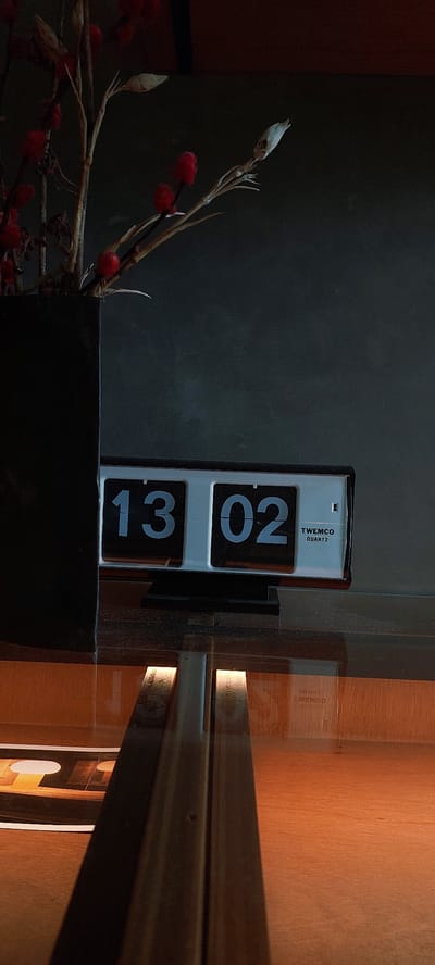 This image shows a close-up of a flip clock displaying "13 02" and a decorative branch with small red pom-poms and dried buds. The clock is white with black numbers and has "TWEMCO QUARTZ" printed on it. It is mounted on a dark gray wall, which contrasts with a wooden surface to the right that is illuminated by warm light, casting long shadows. The lighting suggests it might be late afternoon or early evening. The scene appears to be indoors, possibly in a home or office setting in Sampora, Indonesia, given the provided location context. There are no people visible, and the overall mood is quiet and still, with a focus on the interplay of light, shadow, and the displayed time.