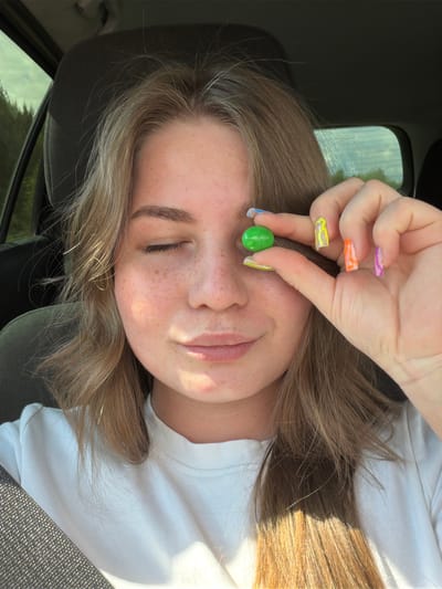 A young woman with blonde hair and freckles is sitting in the driver's seat of a car. She is holding a green candy between her thumb and index finger, bringing it up to her left eye. Her left eye is closed, and she is smiling slightly. Her nails are painted with bright, colorful patterns. The background shows the interior of the car and a glimpse of the outdoors through the windows, suggesting a rural or wooded setting. The location is identified as Oreshniki, Russia.