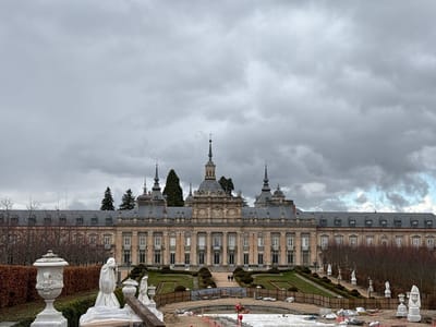 The image presents a wide view of the Royal Palace of La Granja de San Ildefonso and its formal gardens in Real Sitio de San Ildefonso, Spain, under a heavily overcast sky. The multi-story Baroque palace, constructed of light-colored stone, features a symmetrical facade with a prominent central block topped by a domed spire and flanked by smaller spires. Two long, symmetrical wings extend horizontally, each with a grey mansard roof punctuated by dormer windows. Deciduous trees, appearing dormant, line the perimeter of the gardens and background, with a cluster of dark green coniferous trees visible behind the central palace structure.
In the midground, the terraced gardens display manicured green lawns, dark green topiaries, and pathways. Several white classical statues are positioned along these paths, leading towards the palace. Two small human figures are discernible walking on a pathway in the middle ground, approaching the palace entrance.
The immediate foreground reveals an active renovation or construction site. A large, circular basin, presumably a fountain, is empty and partially covered with white tarps, plastic sheeting, and various construction materials including rebar, pipes, and sandbags. White classical urns and sculpted figures, some draped in protective white material, are positioned on a low stone balustrade or wall, with a wooden beam resting upon one of the covered sculptures. Low wooden fencing demarcates parts of the construction area and garden beds.