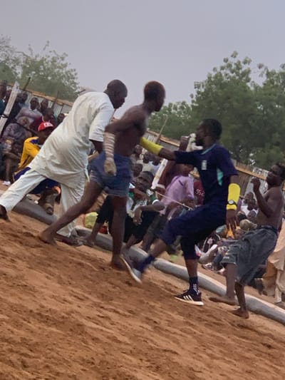 The image shows a group of men engaged in a wrestling match on a sandy field in Sokoto, Nigeria, located at 13.049 latitude and 5.238 longitude. The wrestler on the right is wearing a dark blue shirt with a light blue stripe and yellow wristbands. He is in the middle of a maneuver, his leg outstretched as he makes contact with his opponent. The wrestler on the left is shirtless and wearing blue shorts. A man in a white robe watches the match. There are other onlookers in the background, some sitting and some standing. The scene is set against a backdrop of trees and buildings. The weather appears to be clear, and the ground is a light brown color.