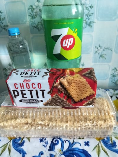 The image shows a collection of food and drink items on a table, likely in a domestic setting in Cupcini, Moldova. In the background, a large green bottle of 7UP soda with the brand's iconic logo is visible. To the left of the soda bottle is a smaller plastic bottle, possibly of water, with a blue cap.
In the foreground, a rectangular box of "Choco Petit" biscuits by "Neris" is prominently displayed. The packaging is red and white, featuring an image of chocolate-covered biscuits. Adjacent to the biscuit box, and slightly behind it, is a clear plastic container holding what appears to be a layered cake or dessert, possibly a napoleon cake, covered in crumbs. The table is covered with a floral patterned tablecloth, featuring blue and green foliage. The overall impression is that of a casual gathering or a snack setup.