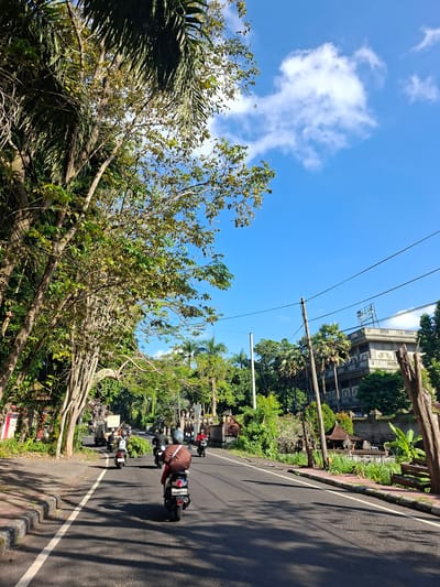 This is a photograph of a sunny day with a bright blue sky and scattered white clouds. The scene is a street in Banjarangkan, Indonesia, lined with lush green trees and vegetation. Several motorbikes are traveling on the paved road, with some riders carrying bundles of green foliage. In the background, a multi-story building with an ornate facade and a sign that reads "MUSEUM" is visible. The overall impression is a vibrant, tropical environment with local transportation and a cultural landmark.