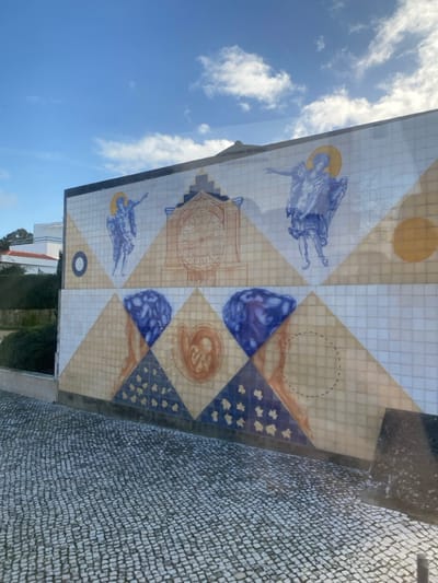 The image presents a detailed view of a large azulejo mural on an exterior wall in Mafra, Portugal. The mural is located on the wall of the Biblioteca Municipal de Mafra, adjacent to the Palácio Nacional de Mafra.
The mural, composed of ceramic tiles, features a complex design laid out in two primary horizontal sections. The foreground consists of a light-colored cobblestone pavement (calçada portuguesa). The sky above the mural is clear blue with scattered white cumulus clouds.
The upper section of the mural includes three distinct elements against a background of alternating white and cream/yellow triangles:
1. On the left, a winged figure, likely an angel or saint, in blue and white, with a golden halo, is depicted gesturing to its right.
2. In the center, an ornate, brown-toned clock face with Roman numerals is prominently displayed. Above the clock, the text "PENSAMENTOS DO REI" is partially visible.
3. On the right, a second winged figure, mirroring the first, also in blue and white with a golden halo, gestures to its left.
The lower section of the mural utilizes a background of white, cream/yellow, and dark blue triangles, some of the dark blue triangles featuring small, light yellow star-like patterns. This section includes:
1. On the far left, a stylized, organic, or abstract form rendered in shades of blue.
2. Next to it, a depiction of a human fetus or embryo, curled within a womb-like structure, rendered in brown and orange tones.
3. Following this, another stylized blue, organic, or abstract form.
4. On the far right, a cream-colored tile section contains a faint, dashed circular outline, indicating an uncompleted or designated area.
To the left of the mural, a low wall separates the pavement from a small garden area with green shrubs and the corner of a white building featuring a terracotta-colored roof. The scene is illuminated by daylight, suggesting a clear day.