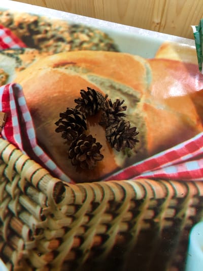 The image shows a close-up view of several pine cones resting on a surface that appears to be a printed mat or tablecloth. The mat has a pattern that resembles a woven basket and a red and white checkered fabric. In the background, there is a large, round object that looks like a loaf of bread, also part of the printed design. The lighting suggests an indoor setting, possibly a table. There is no indication of the city of Izdeshkovo, Russia, or any other specific location in the image.