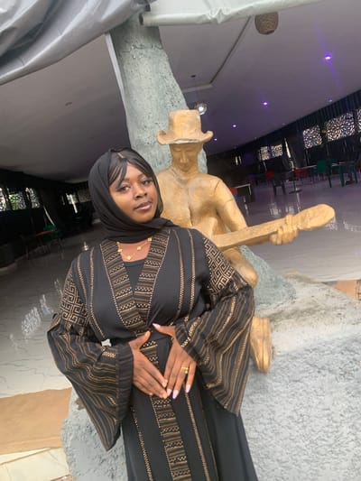 A medium shot captures a Black woman dressed in a black and gold patterned abaya and a black hijab. She is positioned in the foreground, facing the camera with her hands clasped over her stomach. Her gaze is directed slightly to the left of the camera. Behind her, a gold-colored statue of a person wearing a hat and holding a long, flat object is visible, partially obscured by the woman. The statue appears to be seated on a grey, textured surface. The background shows an indoor setting with a tiled floor, tables, and chairs, suggesting a public space such as a restaurant or event hall. The environment is illuminated by overhead lights, some of which cast a purple hue. The overall scene is static, with no apparent interaction or action occurring. The location is specified as being in the city of Bwari, Nigeria.