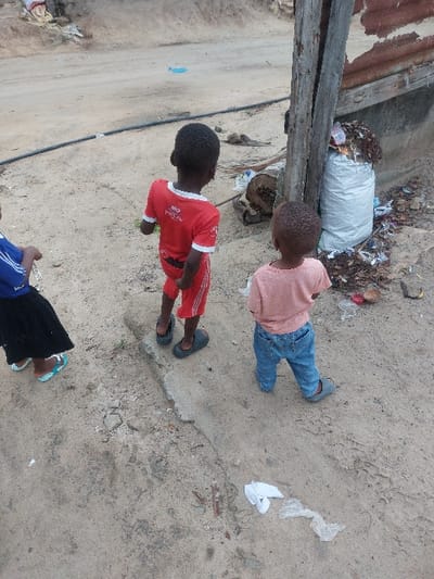An outdoor scene in Mwandege, Tanzania, depicts three children standing on a sandy, unpaved surface next to a dirt road. In the foreground-left, a child, partially visible, wears a blue top, black skirt, and light blue sandals. In the center, a child wears a red t-shirt with white trim and text, red shorts, and dark grey clogs, facing away from the camera towards the road. To their right, a younger child wears a peach t-shirt, blue jeans, and light grey shoes, also facing away from the camera.
To the right of the children, a weathered, vertically oriented wooden post with a visible crack stands next to a white woven sack filled with refuse, including plastic bottles and other debris. Behind the post and sack, a piece of corrugated metal is visible. Scattered refuse, including plastic and organic material, lies on the ground around the post and sack. A black pipe runs horizontally across the mid-ground, traversing the sandy terrain. The background features a wider dirt road with visible tracks, and distant, indistinct structures or mounds of earth. The ground is primarily dry earth and sand, with scattered small pieces of white plastic and other debris in the foreground.