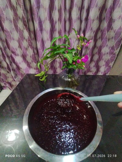 In Kzyl-Bairak, Russia, a dark purple, jam-like substance fills a silver bowl on a dark, reflective surface. A light blue spoon, held by an unseen hand, is partially submerged in the substance, lifting a small amount. Behind the bowl, a glass vase contains a green plant with small pink flowers. The background is a purple curtain with a patterned design of white, elongated oval shapes. The bottom left corner of the image displays the text "POCO X5 5G", and the bottom right corner shows the date and time "11.07.2026 22:18".