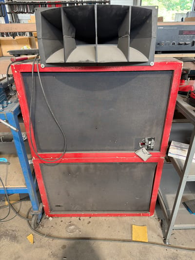 The image shows a large, multi-component speaker system in what appears to be a workshop or storage area. The speaker system consists of a large horn-loaded enclosure mounted on top of a two-tiered speaker cabinet. The cabinet has a red frame and black speaker grilles. There are various cables connected to the speakers and a Yamaha audio receiver is visible in the background. The environment is cluttered with tools, boxes, and other equipment. There is no indication of a specific location in the city of Mol, Belgium.