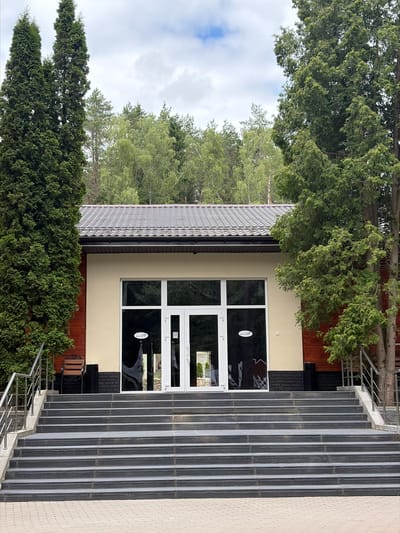 The image depicts the exterior of a building with a prominent entrance. A large set of stairs with metal railings leads up to double glass doors. The building has light-colored walls and a dark, textured roof. Adjacent to the entrance, there are large windows, one of which displays a logo with text. The building is surrounded by lush green trees and foliage, suggesting a natural or park-like setting. The sky is partly cloudy, indicating daytime. The scene appears to be a quiet, possibly public, establishment. The text on the logo is not clearly legible. The location is identified as Piatryshkaŭski Sel'ski Savet, Belarus.