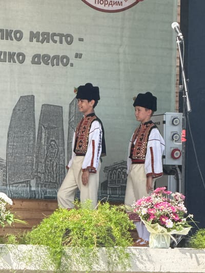 Two young boys, dressed in traditional Bulgarian folk costumes, stand on a wooden stage during an outdoor event in Pordim, Bulgaria. The boy on the left wears a black hat with a red flower, a white shirt with embroidered sleeves, and a decorative vest. The boy on the right, also wearing a black hat, a white shirt with embroidered sleeves, and a vest, stands slightly behind him. Behind them, a large banner displays an image of a monument and text in Bulgarian. A microphone on a stand is visible on the right side of the stage. In front of the stage, a bouquet of pink and white flowers sits on a ledge, flanked by lush green foliage. The overall scene suggests a cultural performance or celebration.