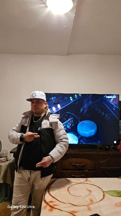 This image, taken in Real Sitio de San Ildefonso, Spain, depicts a male individual standing indoors, positioned slightly off-center to the left, facing the camera. The man is wearing a white baseball cap, a silver chain, a black t-shirt, a light grey puffer jacket with black shoulder and chest panels, and light-colored sweatpants. A black smartwatch is visible on his left wrist. His hands are raised to chest level, both forming a "peace sign" or two-finger gesture.
Behind the man, a large flat-screen television displays an image of illuminated electronic equipment, possibly a music mixing console, featuring various knobs and a prominent bright blue circular element. The word "squirrel" is partially visible in the top right corner of the TV screen. The television rests on a dark wooden cabinet with two drawers. To the left of the cabinet and partially obscured by the man, a small table draped with a patterned cloth holds a fan and a lamp with a spherical glass shade.
In the foreground, to the right, a light-colored blanket with a distinct brown and orange swirl and leaf pattern covers a surface, likely a bed or sofa. A bright circular ceiling light fixture is visible at the top center of the image, emitting a strong glare. The room walls are plain and light-colored. The bottom left corner of the image contains a "Galaxy S24 Ultra" watermark.