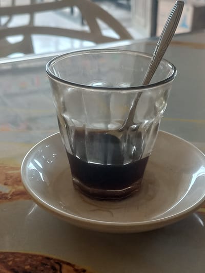 A close-up, eye-level shot captures a small, clear glass filled with dark liquid, presumably coffee, resting on a white saucer. A metal spoon with a decorative pattern is partially submerged in the liquid, angled towards the right side of the glass. The glass exhibits some condensation on its exterior, and the liquid level is about one-third of the way up the glass. The saucer has a slightly raised rim and appears to be made of ceramic. The surface beneath the saucer has a textured, patterned design in shades of brown and beige. In the background, blurred out, are suggestions of an indoor setting with light-colored furniture and a window. The overall lighting is soft and natural.