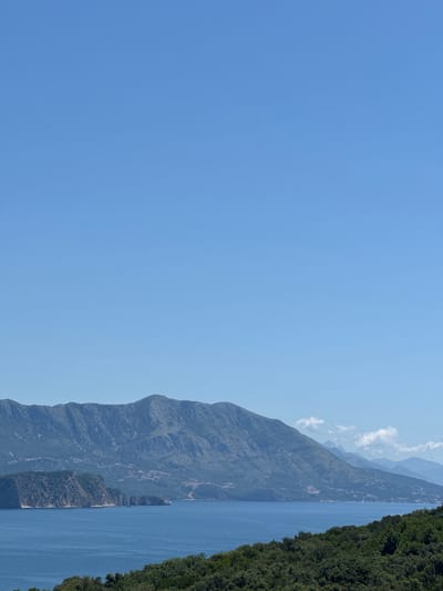 This wide-angle daytime image captures a vast coastal landscape in Kubasi, Montenegro, under a clear azure sky that occupies approximately the top two-thirds of the frame. The horizon line is dominated by a rugged, multi-layered mountain range, appearing dark green to bluish-gray, with settlements visible as small clusters of structures on their lower slopes facing the sea. In the midground, a significant body of tranquil, deep blue water stretches across the scene. A prominent, verdant peninsula or large island with a steep, rocky cliff face extends from the left side into the water. The immediate foreground is densely covered by dark green foliage, suggesting an elevated viewpoint overlooking the coastline. No people, specific objects (beyond natural features and distant buildings), actions, or interactions are discernible within the frame.