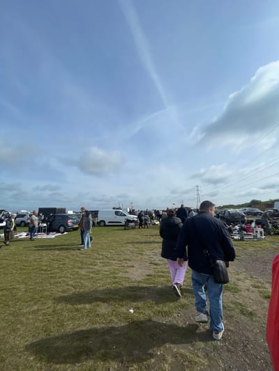 The image depicts an outdoor marketplace in Sevenoaks, United Kingdom, under a cloudy sky. A line of parked vehicles is visible along a grassy field. Numerous individuals are present, some inspecting goods laid out on blankets or tables near the cars. Overhead power lines are also visible. The focus is on the attendees and their activities, set against the backdrop of the field and parked vehicles.