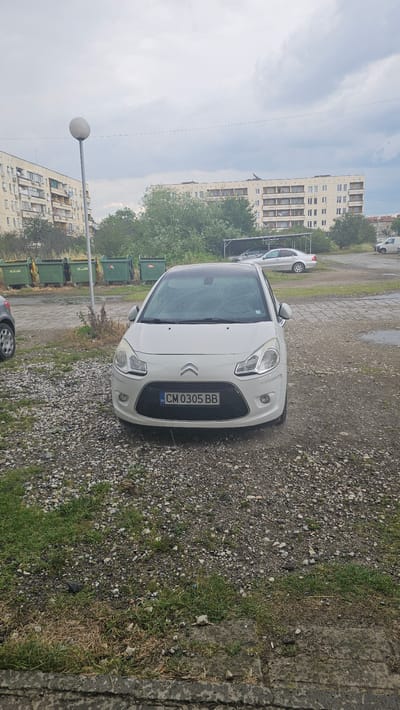 A white Citroën C3 is parked on a gravel lot in front of a row of green dumpsters, with residential buildings and trees in the background. The license plate reads "CM 0305 BB". The sky is overcast, and the ground appears wet, suggesting recent rain. The scene depicts an urban residential area, likely in Vedrare, Bulgaria, given the license plate format.