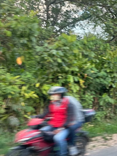 The image shows a motorcycle with a rider on a road adjacent to foliage in Dak Ro Wa, Vietnam. The rider is wearing a helmet, red jacket and blue jeans. The motorcycle is mostly red and black. The road surface is light gray and appears to be paved. There are trees and dense green vegetation nearby. The image is blurred, suggesting motion.